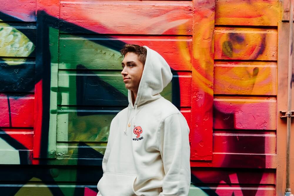 Pria mengenakan hoodie berwarna putih, berdiri di depan tembok yang digambari grafiti.